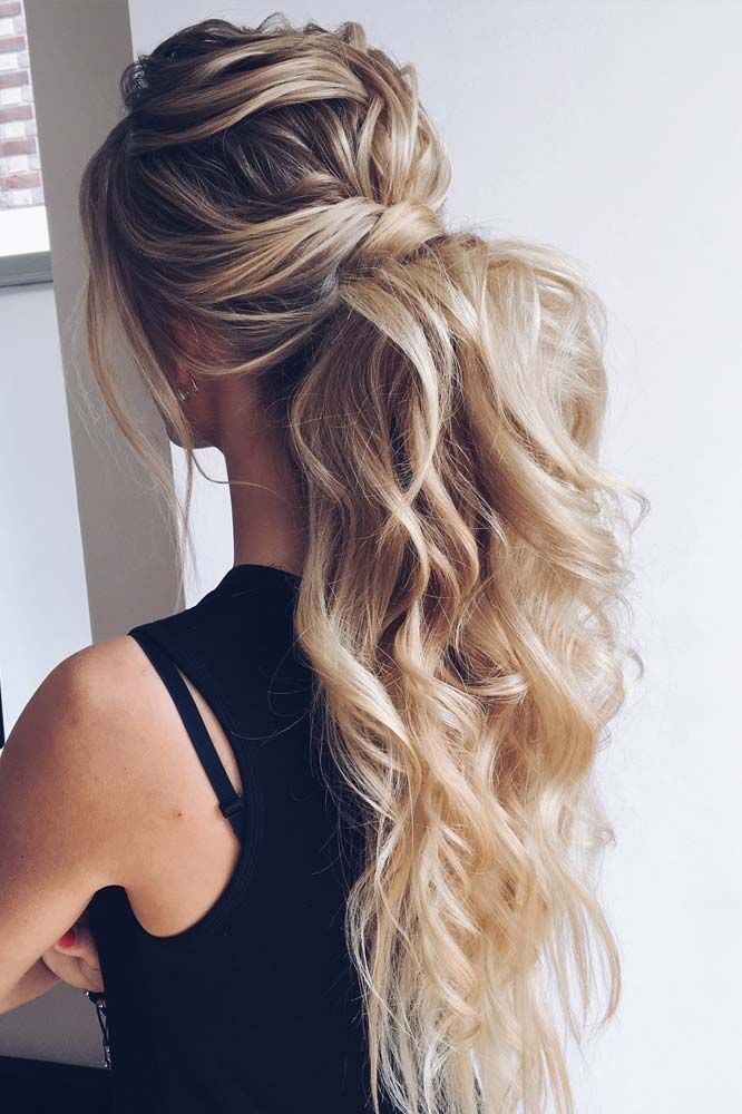 Back view of a woman with long wavy blonde hair styled in a half-up ponytail, wearing a black top near a window.