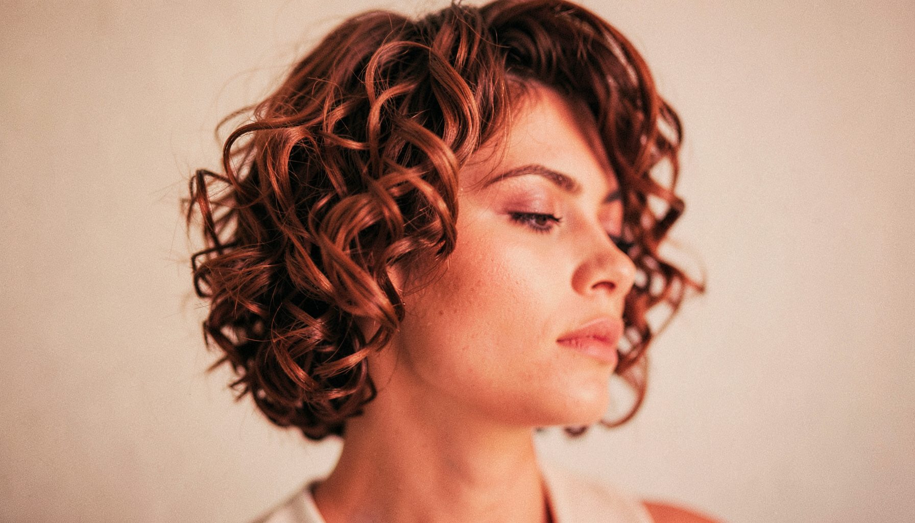 Soft warm-toned editorial shot of a short curly hairstyle with bouncy ringlets and a side part, shallow depth of field