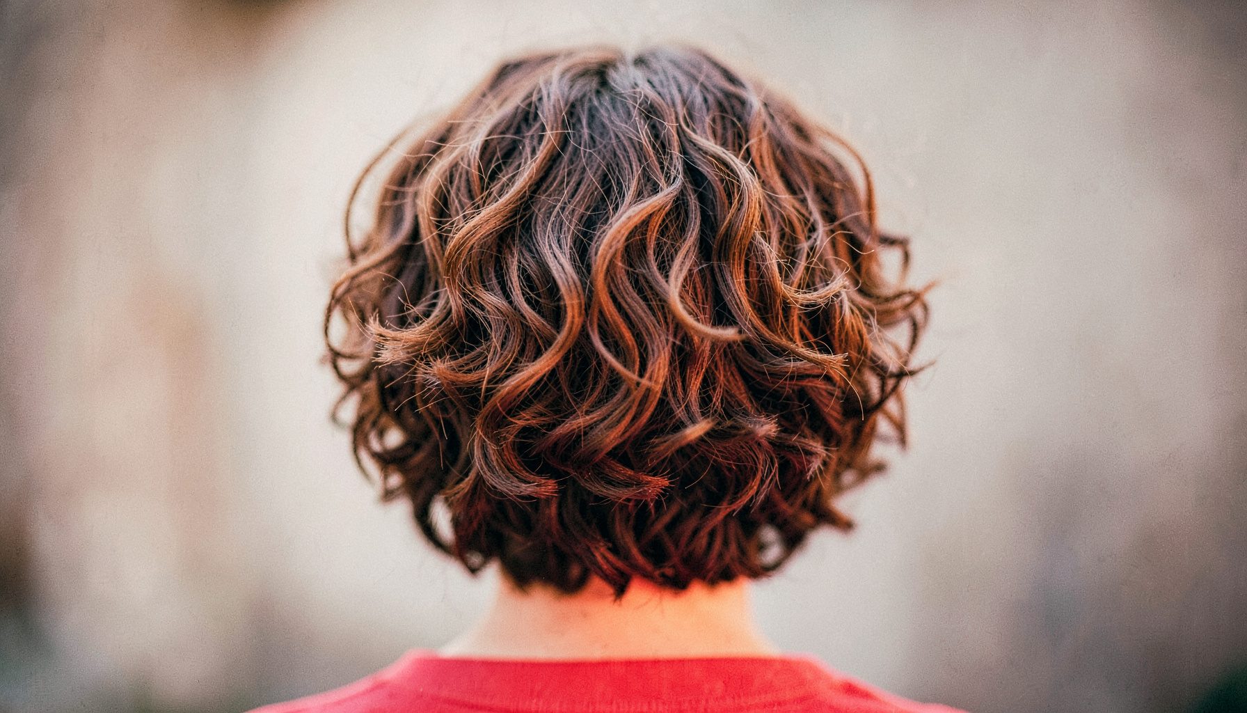 Back view of a layered short curly hairstyle in soft brown tones against a blurred neutral background, shallow depth of field