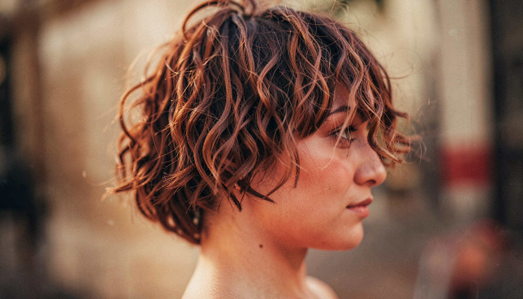 Side profile of a textured short crop with curls and a wispy fringe, muted warm palette, shallow depth of field